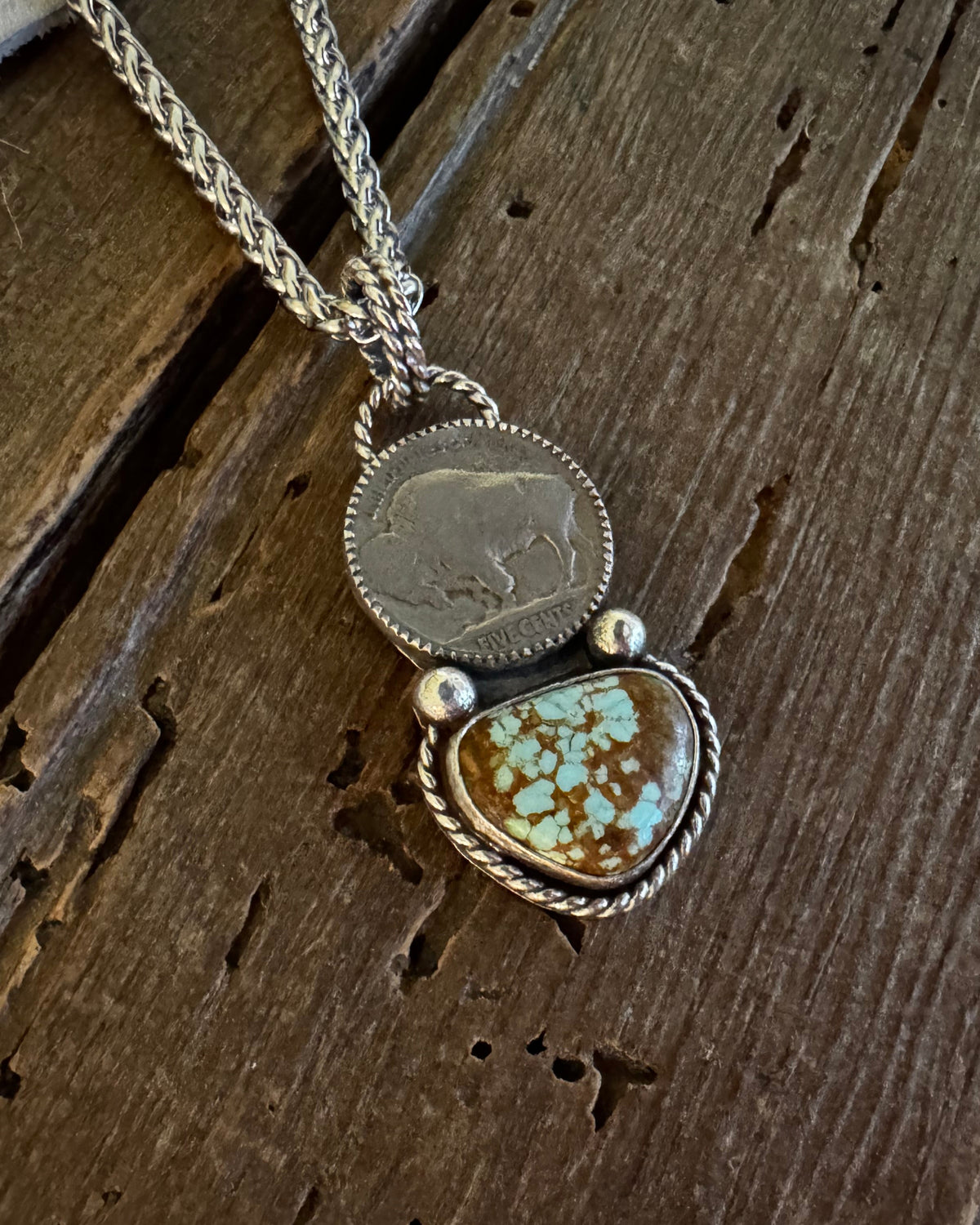 A silver-toned pendant necklace featuring a round coin-style charm and a larger heart-shaped stone pendant with turquoise-like blue and brown marbling, displayed on a rustic dark wood surface.