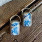 A pair of rectangular drop earrings with a blue and white marbled stone inlay and gold-tone metal borders, displayed on a rustic dark wood surface.