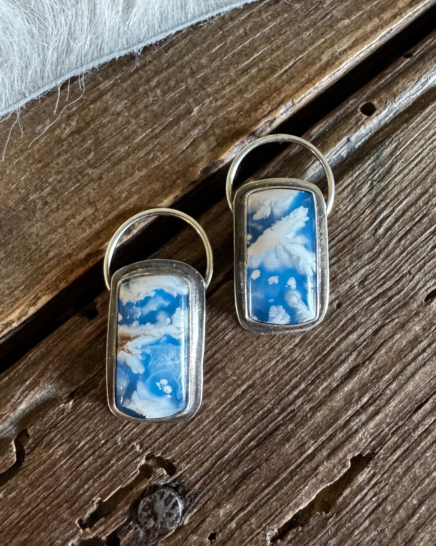 A pair of rectangular drop earrings with a blue and white marbled stone inlay and gold-tone metal borders, displayed on a rustic dark wood surface.