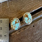 A pair of oval turquoise stud earrings with gold-toned bezel settings, displayed on a rustic wooden surface next to a metal ruler for scale.