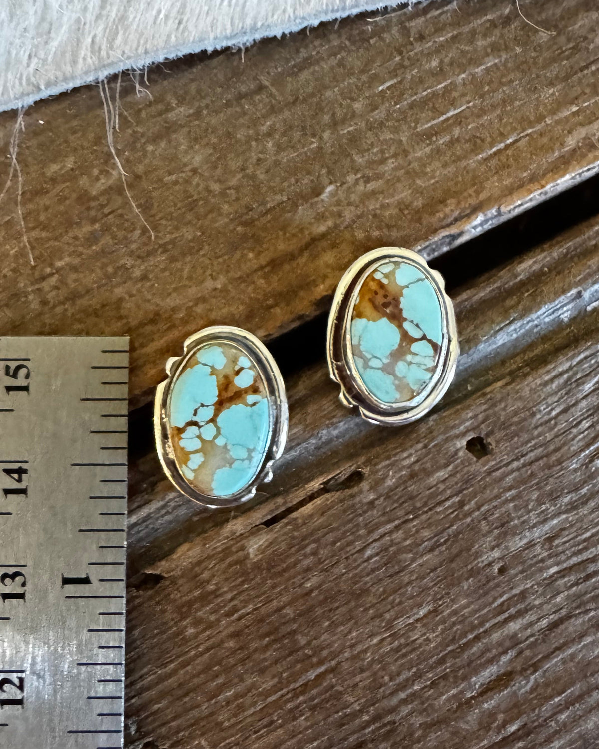A pair of oval turquoise stud earrings with gold-toned bezel settings, displayed on a rustic wooden surface next to a metal ruler for scale.