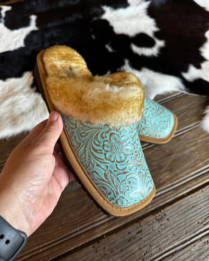 A hand holds a turquoise tooled leather slipper with a tan faux-fur collar, set against a rustic wood floor and a black-and-white cowhide rug.