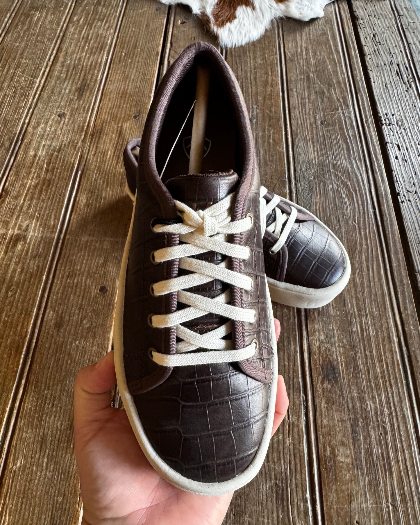 Brown leather sneaker with white laces held by a hand on a wooden surface