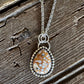 A silver-toned teardrop pendant necklace with a mottled orange, white, and brown stone, displayed on a rustic, weathered wooden surface.