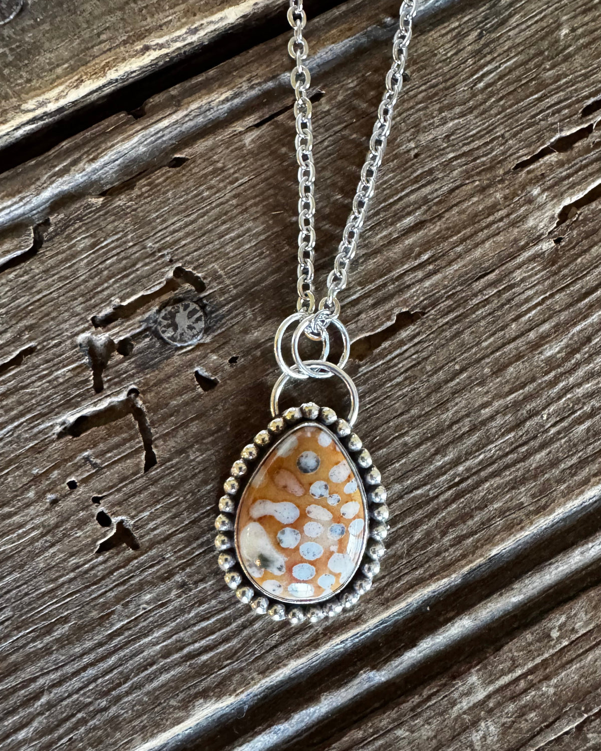 A silver-toned teardrop pendant necklace with a mottled orange, white, and brown stone, displayed on a rustic, weathered wooden surface.