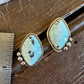 A pair of turquoise-blue stone stud earrings with gold-toned settings and small bead accents, placed on a rustic wooden surface next to a metal ruler for scale.