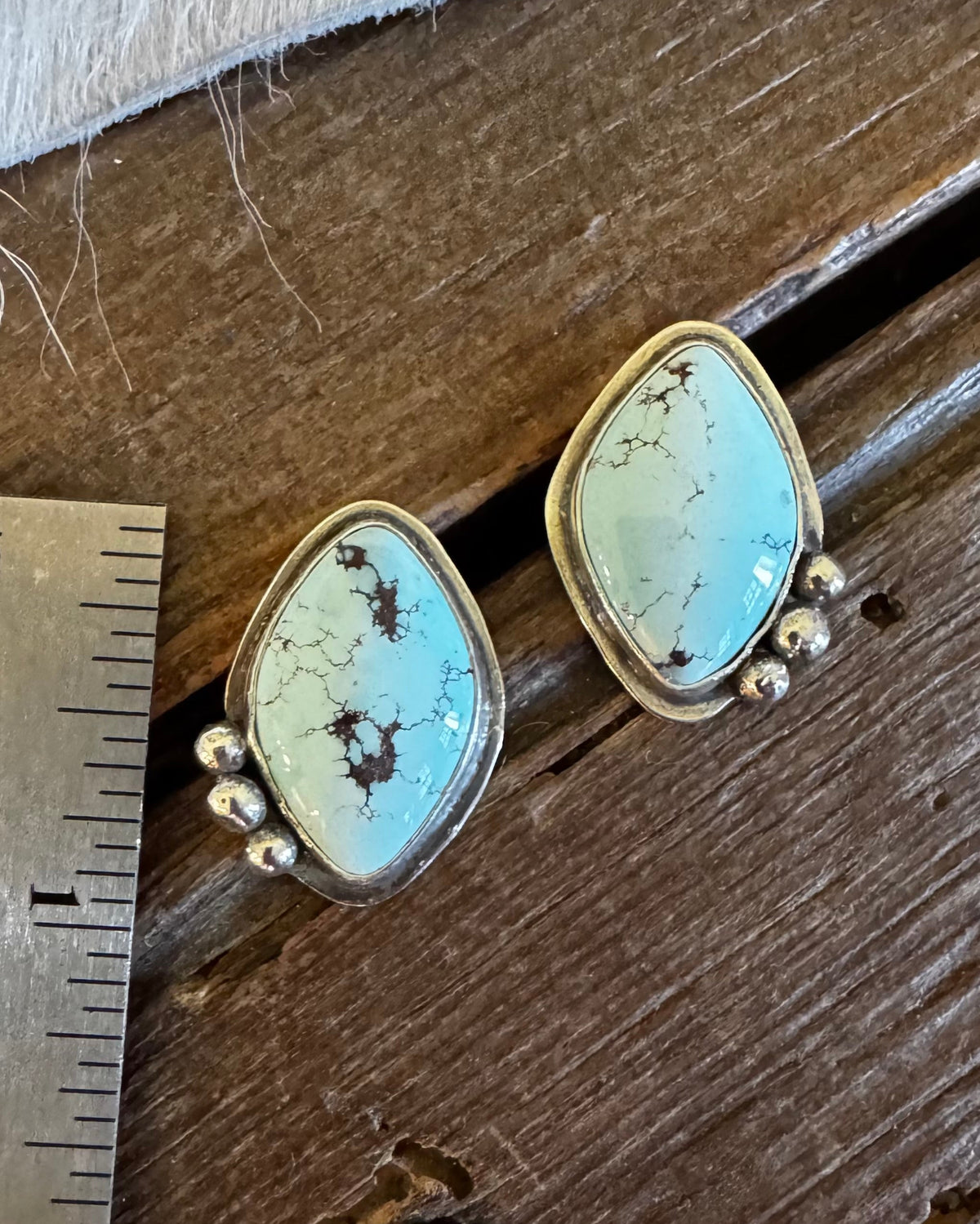 A pair of turquoise-blue stone stud earrings with gold-toned settings and small bead accents, placed on a rustic wooden surface next to a metal ruler for scale.