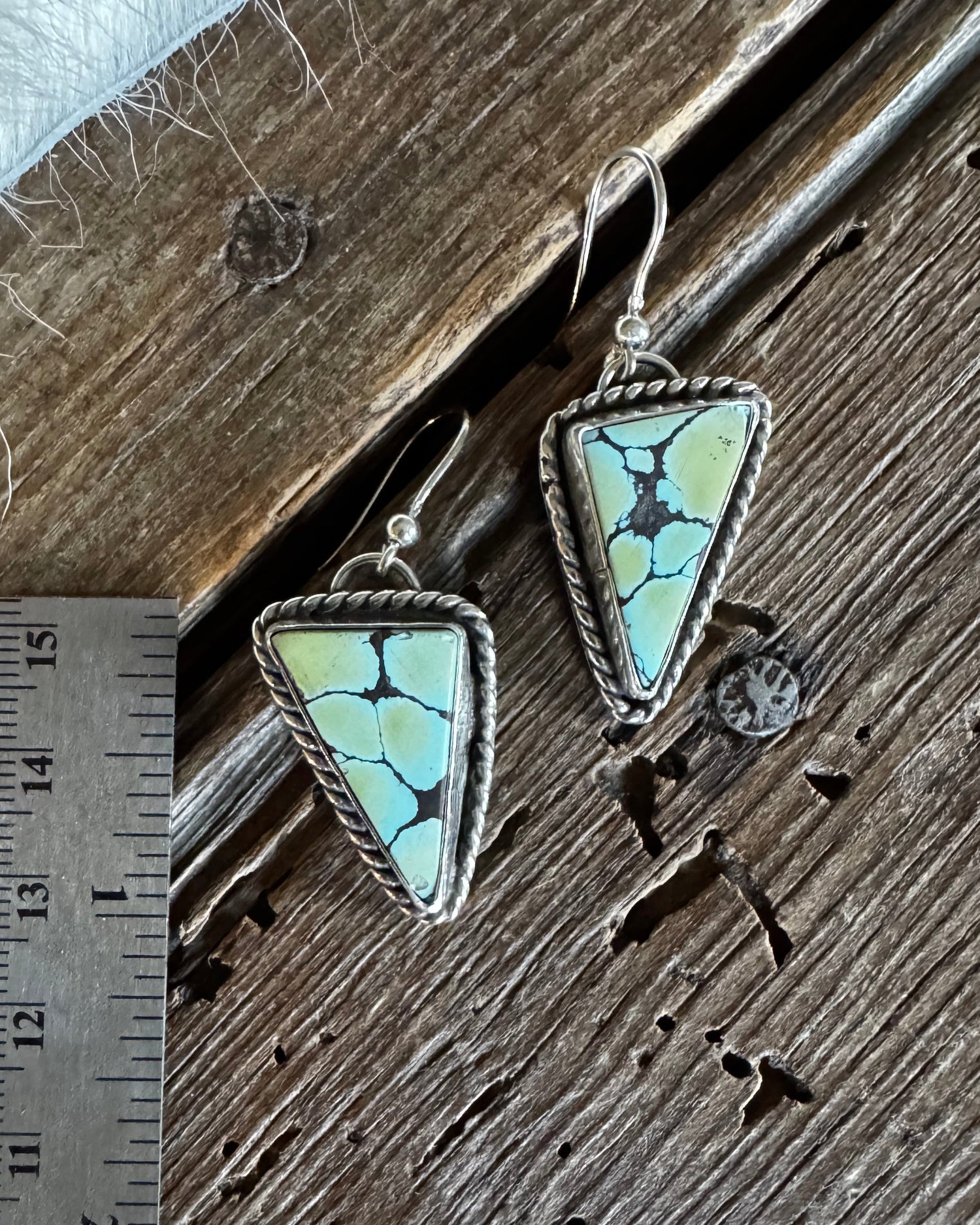 Two triangular turquoise drop earrings with silver bezel settings and fishhook wires, displayed on a rustic wooden surface next to a metal ruler for scale.
