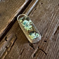 A small oval pendant or charm with a green and white marbled pattern inside a clear resin or glass case, resting on a weathered wooden surface next to white fur.