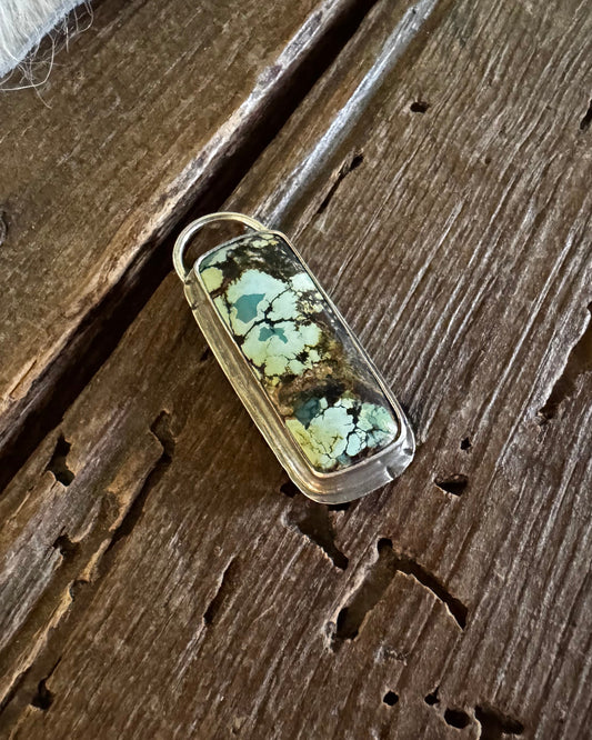 A small oval pendant or charm with a green and white marbled pattern inside a clear resin or glass case, resting on a weathered wooden surface next to white fur.