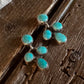 A pair of gold-toned earrings with multiple oval turquoise stones, displayed on a rustic dark wood surface with a white fur trim visible in the top left corner.