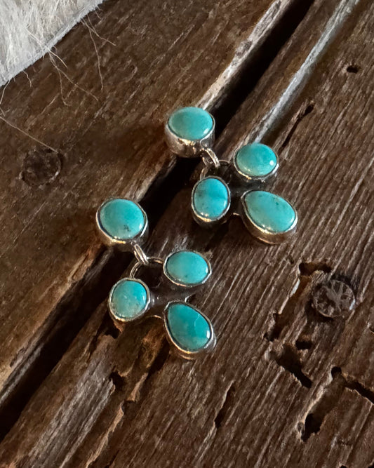 A pair of gold-toned earrings with multiple oval turquoise stones, displayed on a rustic dark wood surface with a white fur trim visible in the top left corner.