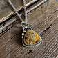 A silver-toned necklace with a triangular pendant featuring a yellow and white marbled stone, displayed on a rustic dark wood surface.