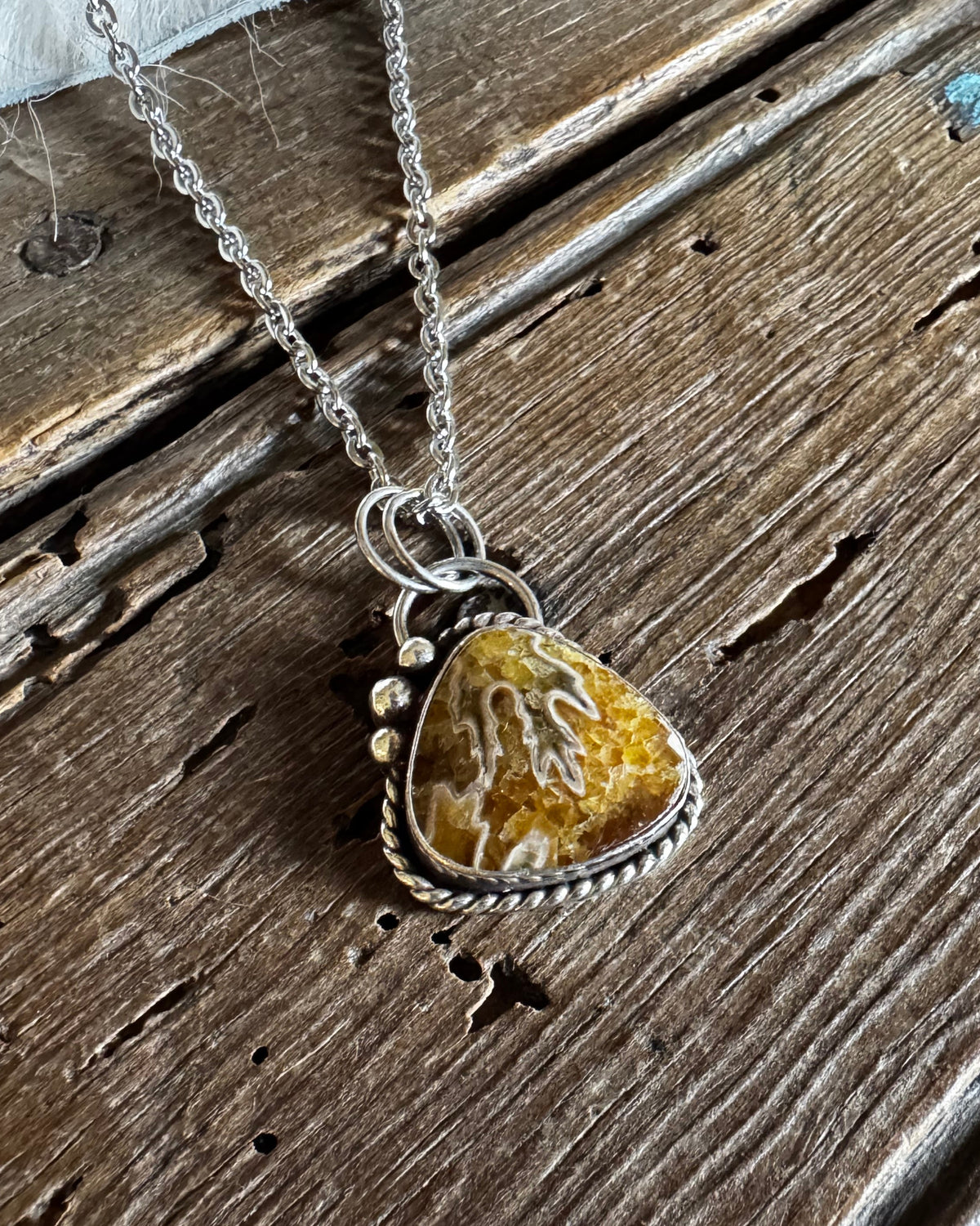 A silver-toned necklace with a triangular pendant featuring a yellow and white marbled stone, displayed on a rustic dark wood surface.