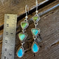 A pair of silver drop earrings with green and turquoise stones, shown next to a metal ruler on a rustic wooden surface.