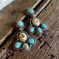 A pair of silver-toned dangle earrings with turquoise and orange-brown stone accents, displayed on a rustic wooden surface next to a white faux-fur textile.