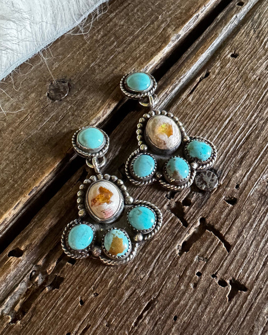 A pair of silver-toned dangle earrings with turquoise and orange-brown stone accents, displayed on a rustic wooden surface next to a white faux-fur textile.