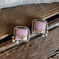 A pair of square stud earrings with pink stone centers and twisted silver-tone borders, resting on a rustic wooden surface with white faux fur above.