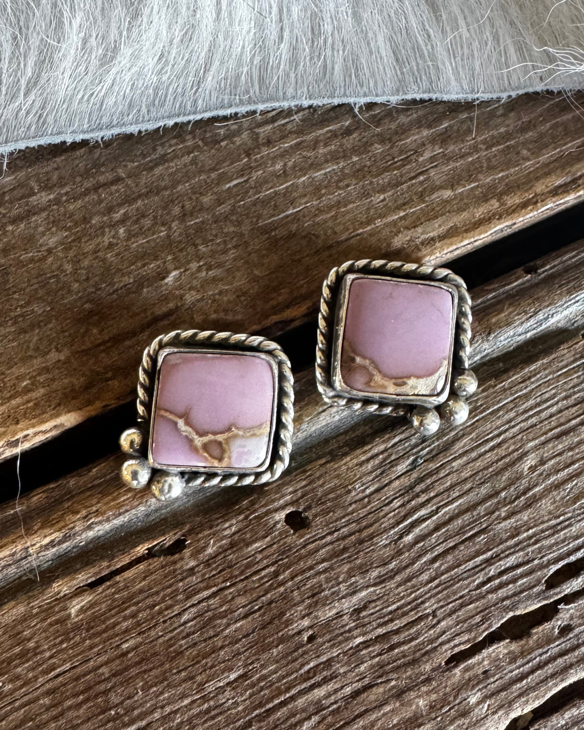 A pair of square stud earrings with pink stone centers and twisted silver-tone borders, resting on a rustic wooden surface with white faux fur above.