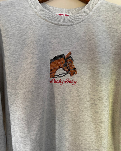 A light heather gray crewneck sweatshirt featuring a pixelated brown horse graphic and the text 'Lucky Baby' embroidered in red on the chest.