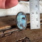 A hand holds a silver-toned ring with an oval turquoise-blue stone next to a stainless steel ruler for scale, set on a rustic wooden surface.