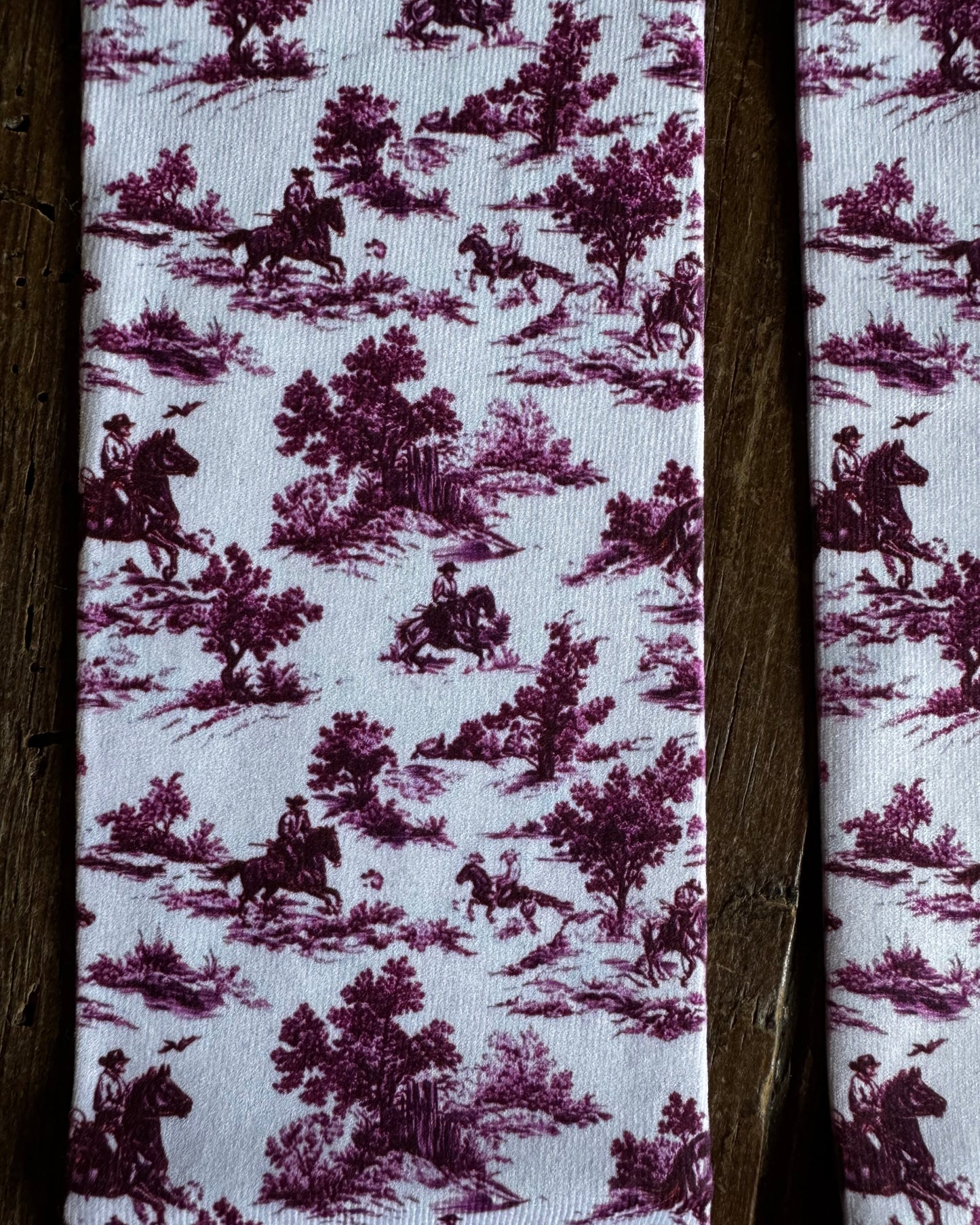A close-up of a fabric product featuring a classic purple and white toile de Jouy pattern with riders and trees, framed by dark brown wooden slats.