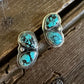 A pair of silver stud earrings with two oval turquoise stones each, displayed on a rustic dark wood surface next to a white textured fabric.