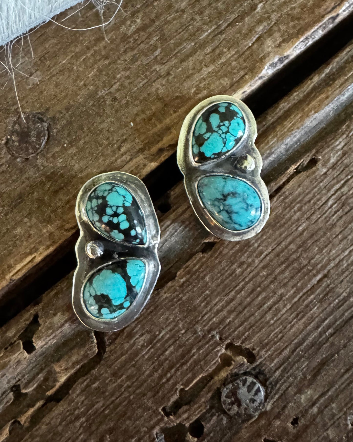 A pair of silver stud earrings with two oval turquoise stones each, displayed on a rustic dark wood surface next to a white textured fabric.