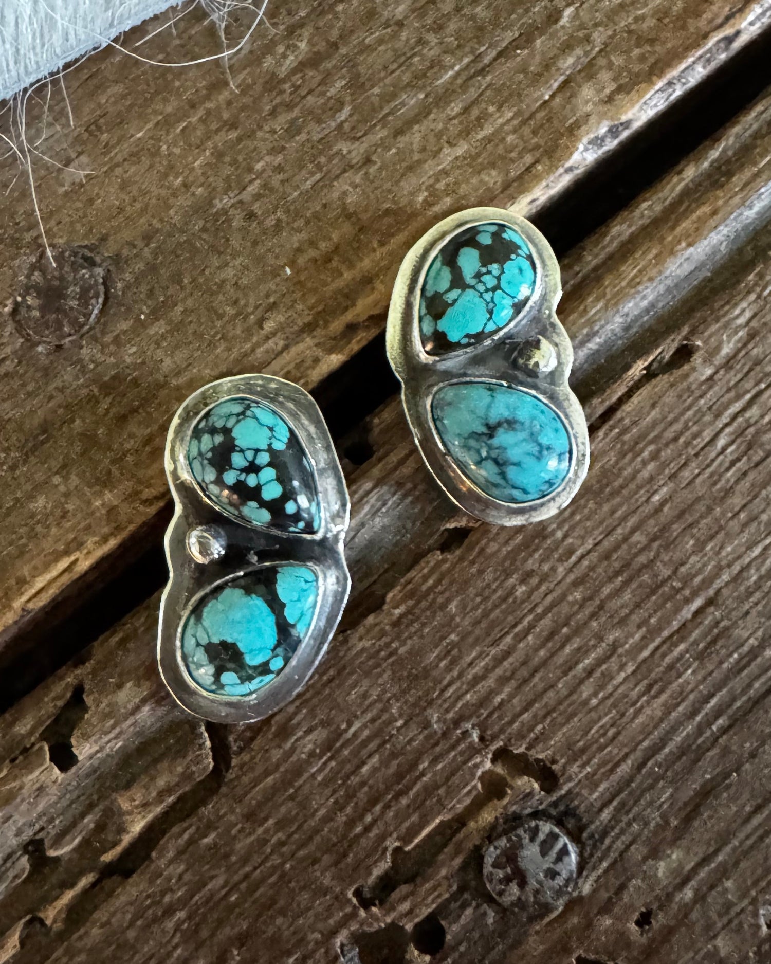 A pair of silver stud earrings with two oval turquoise stones each, displayed on a rustic dark wood surface next to a white textured fabric.