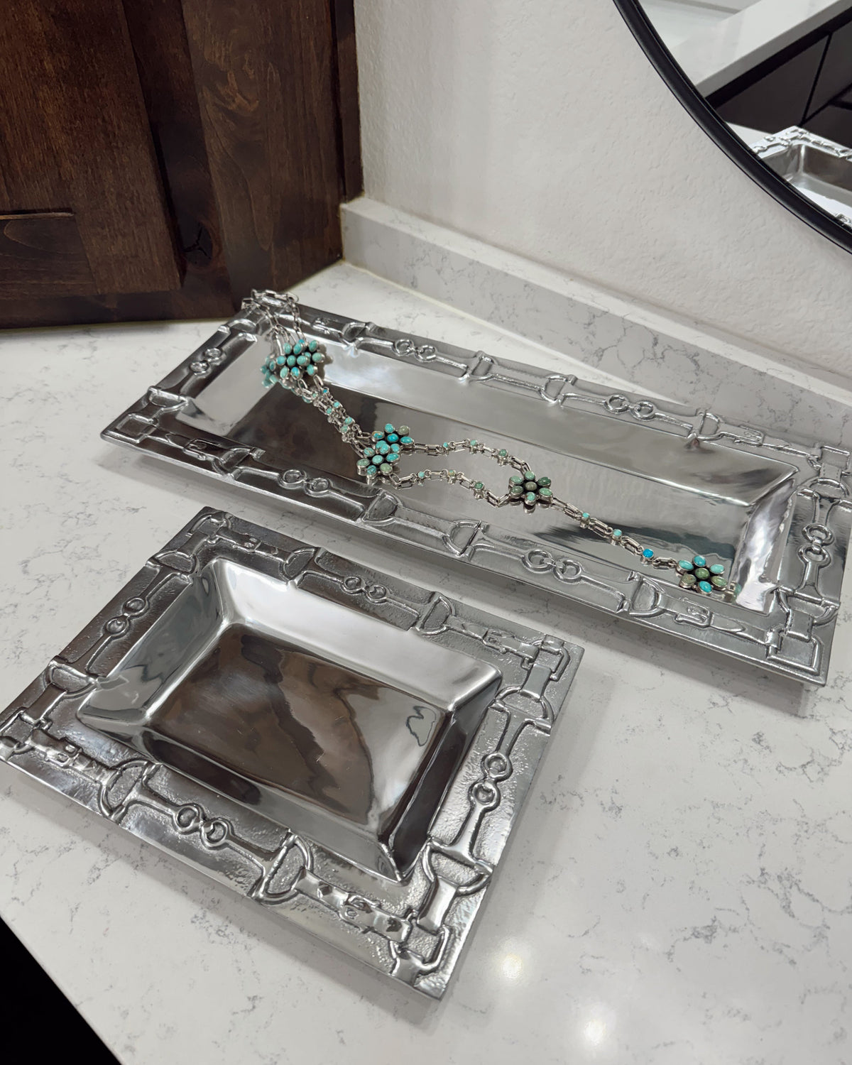 Two silver jewelry trays on a marble surface with a wooden cabinet in the background.