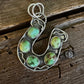 A silver horseshoe-shaped pendant featuring five round green turquoise stones, displayed on a rustic dark wood background.