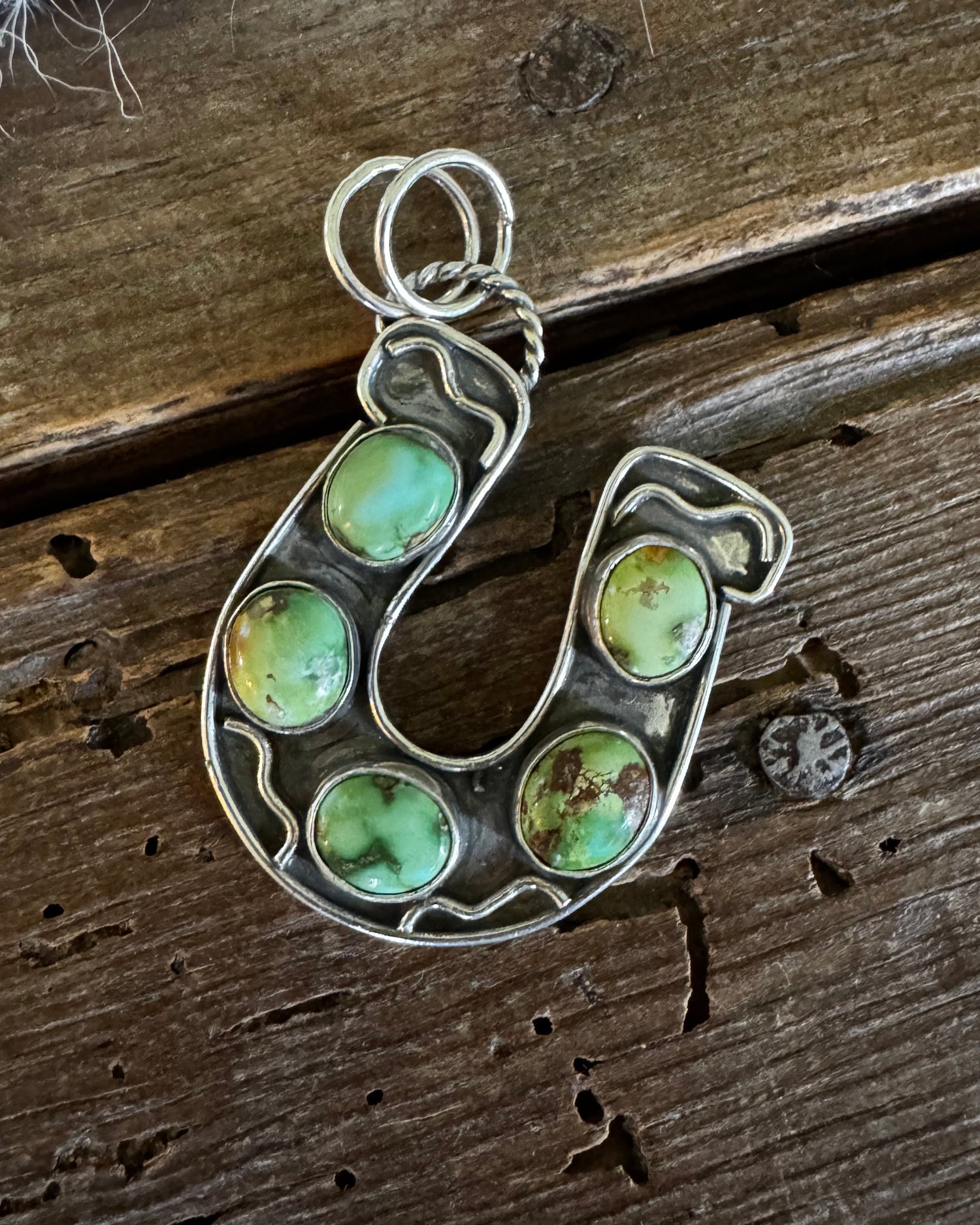 A silver horseshoe-shaped pendant featuring five round green turquoise stones, displayed on a rustic dark wood background.