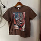 A brown short-sleeve crop top t-shirt with a colorful graphic print of a cowboy boot, hanging on a wooden hanger against a white wall.
