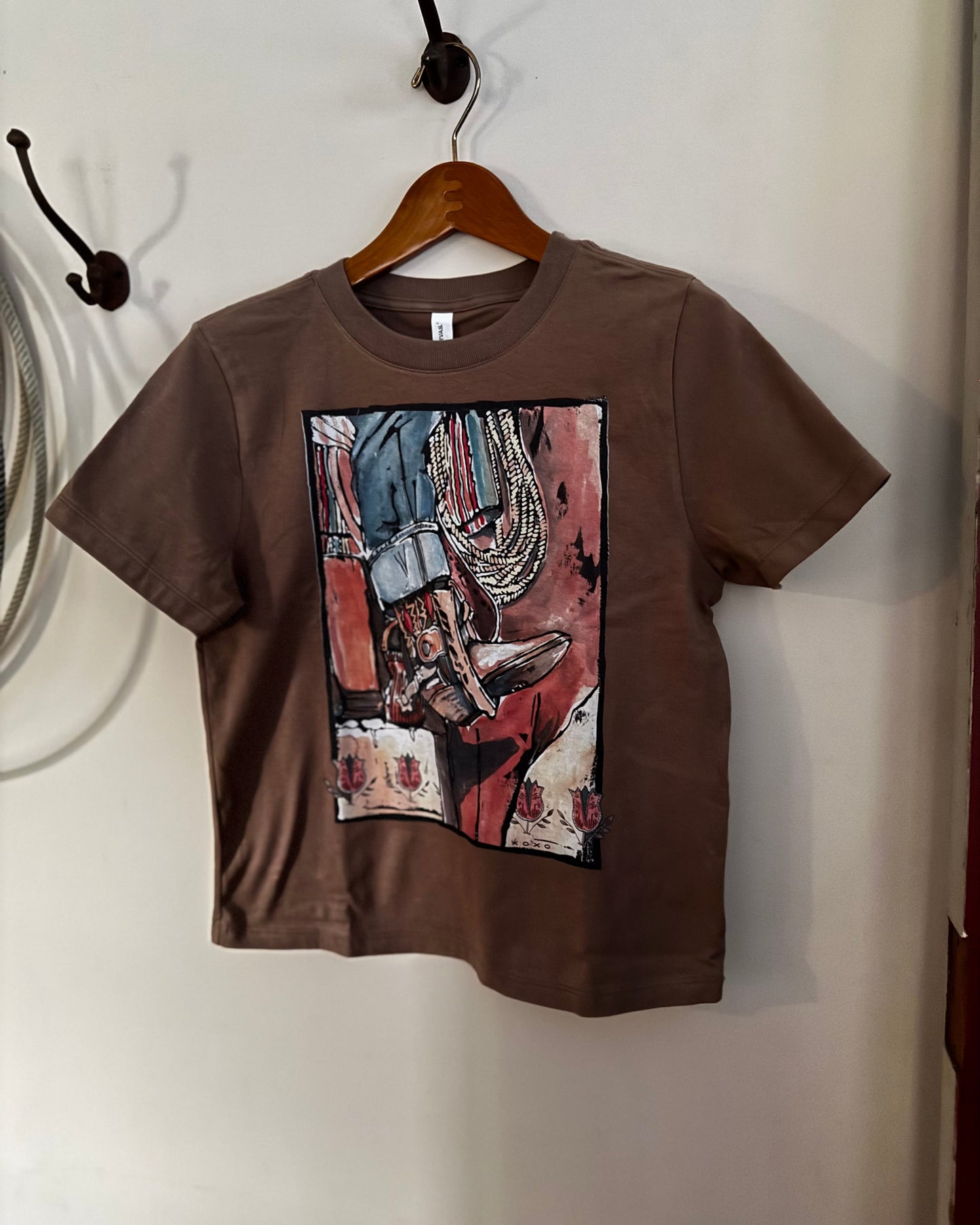 A brown short-sleeve crop top t-shirt with a colorful graphic print of a cowboy boot, hanging on a wooden hanger against a white wall.