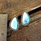A pair of teardrop-shaped turquoise stud earrings with silver bezel settings, displayed on a rustic wooden surface next to a metal ruler for scale.