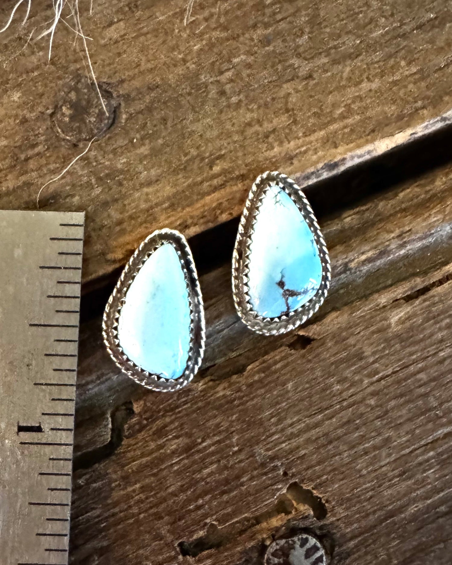 A pair of teardrop-shaped turquoise stud earrings with silver bezel settings, displayed on a rustic wooden surface next to a metal ruler for scale.