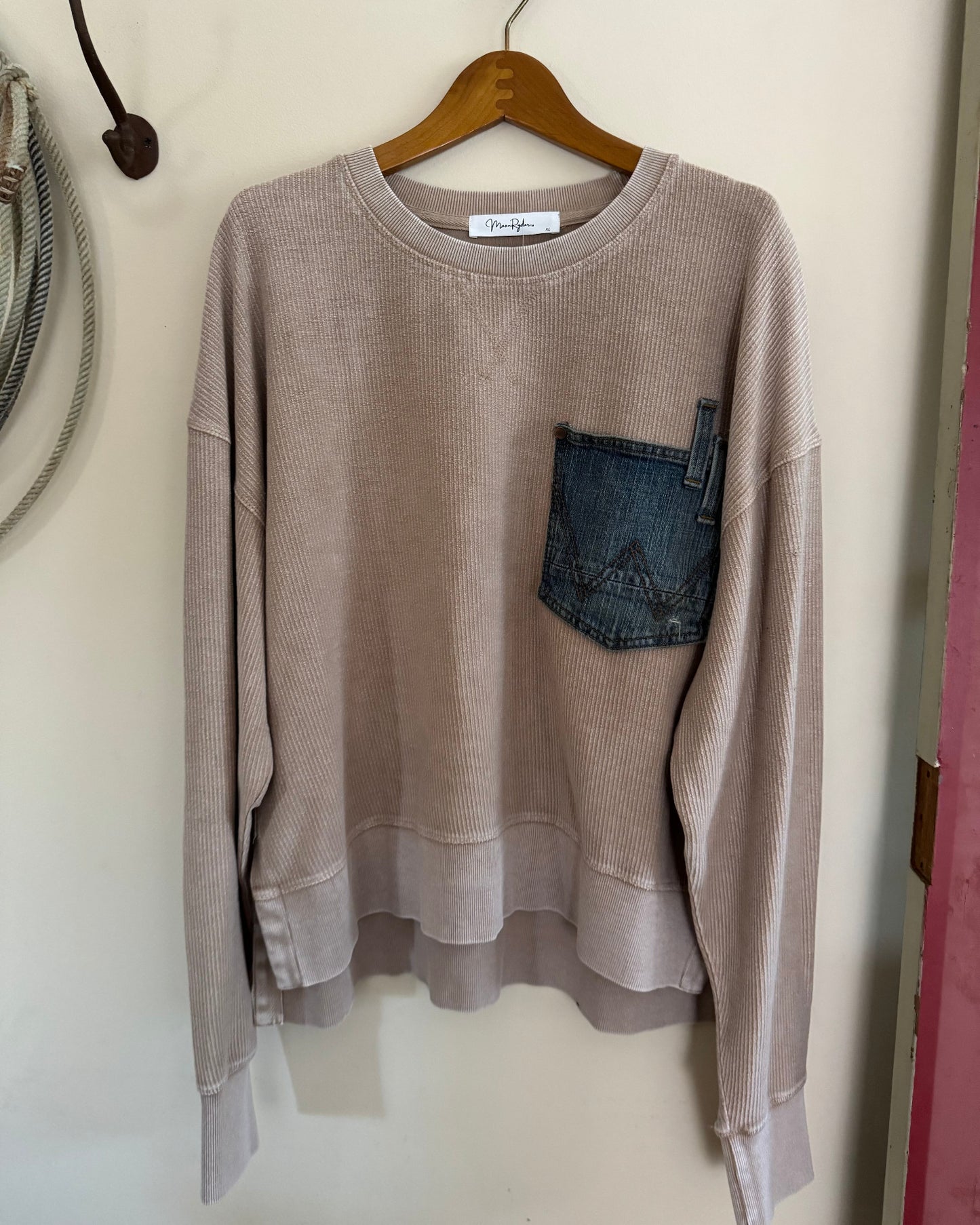Beige sweatshirt with denim pocket on a hanger against a white wall