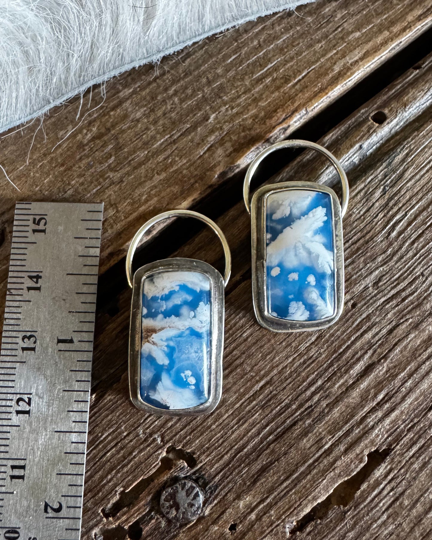Two matching rectangular pendant earrings with blue and white marbled inlay and silver-tone metal frames, displayed on a rustic wooden surface next to a metal ruler for scale.