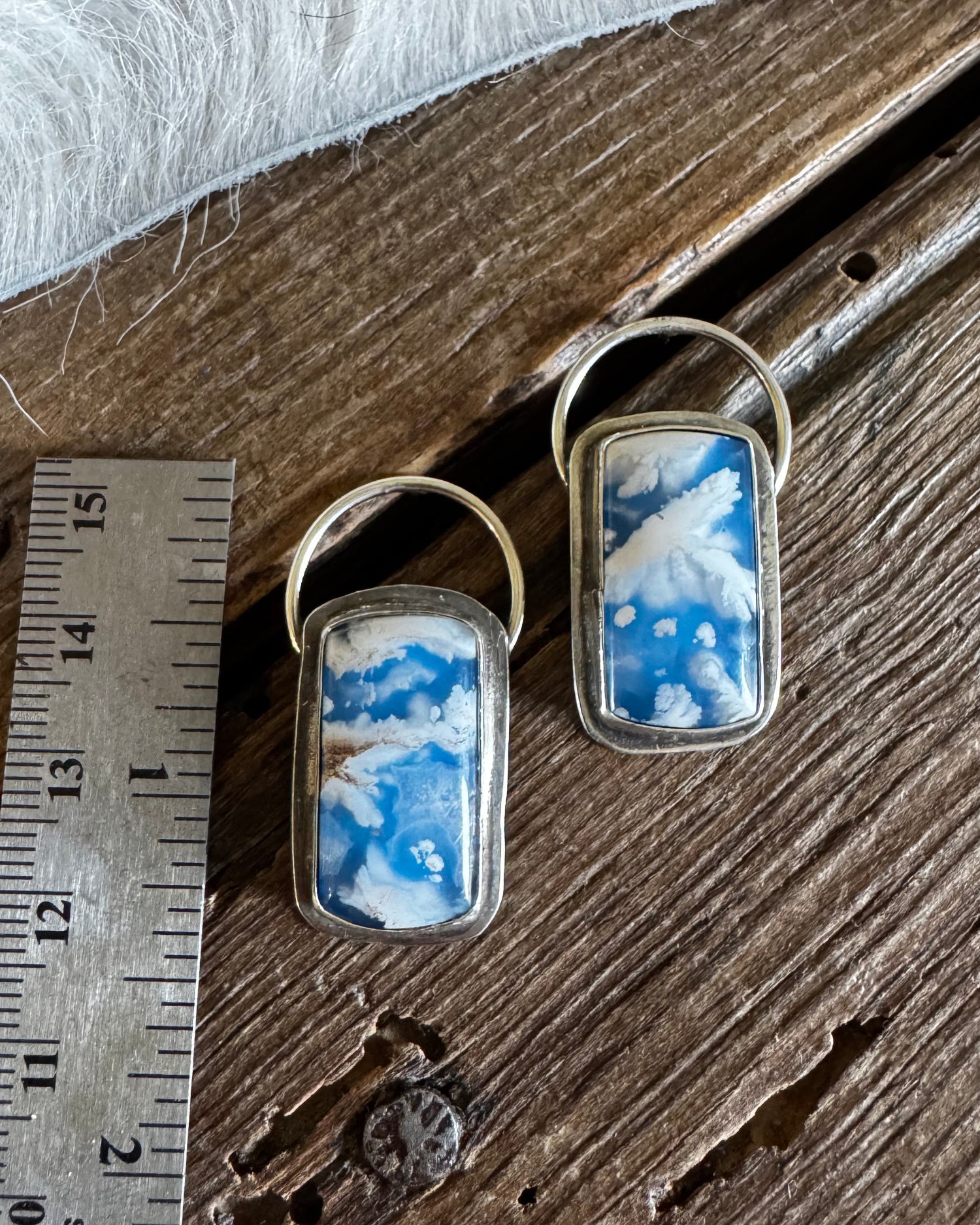 Two matching rectangular pendant earrings with blue and white marbled inlay and silver-tone metal frames, displayed on a rustic wooden surface next to a metal ruler for scale.