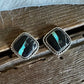 A pair of square stud earrings with black stone inlays and bright turquoise accents, set in a braided silver-tone border, resting on a rustic wooden surface with white faux fur above.