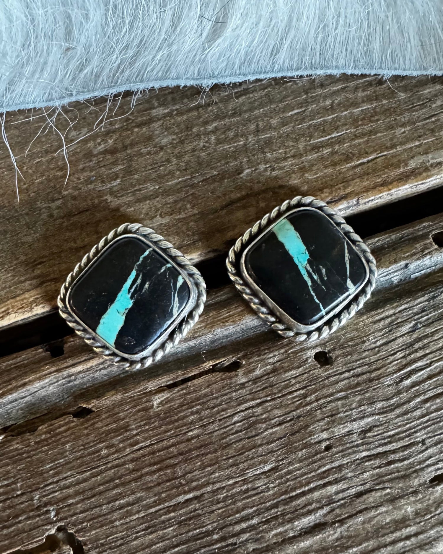 A pair of square stud earrings with black stone inlays and bright turquoise accents, set in a braided silver-tone border, resting on a rustic wooden surface with white faux fur above.
