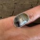 A silver ring with a large, polished cat's eye stone worn on a person's finger, set against a rustic wooden background.