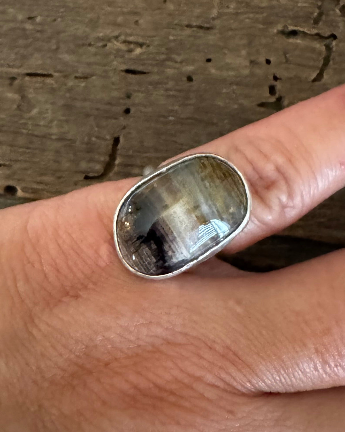 A silver ring with a large, polished cat's eye stone worn on a person's finger, set against a rustic wooden background.