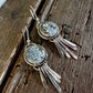 A pair of silver-toned dangle earrings with round textured gray stone centers and dangling leaf-like metal elements, displayed on a rustic dark wood surface.