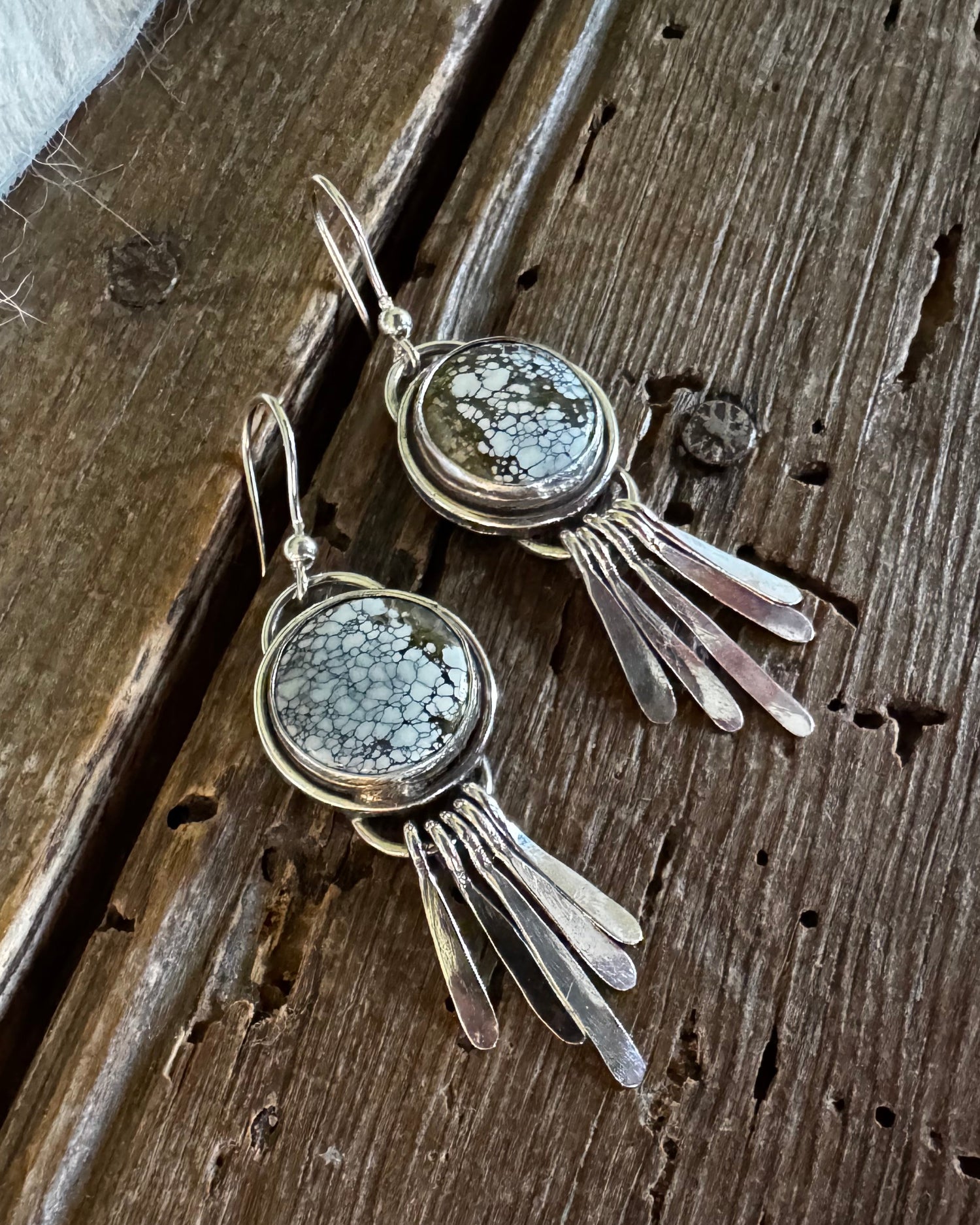 A pair of silver-toned dangle earrings with round textured gray stone centers and dangling leaf-like metal elements, displayed on a rustic dark wood surface.