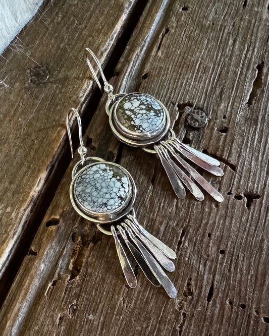 A pair of silver-toned dangle earrings with round textured gray stone centers and dangling leaf-like metal elements, displayed on a rustic dark wood surface.