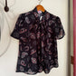 A short-sleeve black blouse with a paisley and hat pattern in pink, white, and red, hanging on a wooden hanger from a metal hook on a white wall.