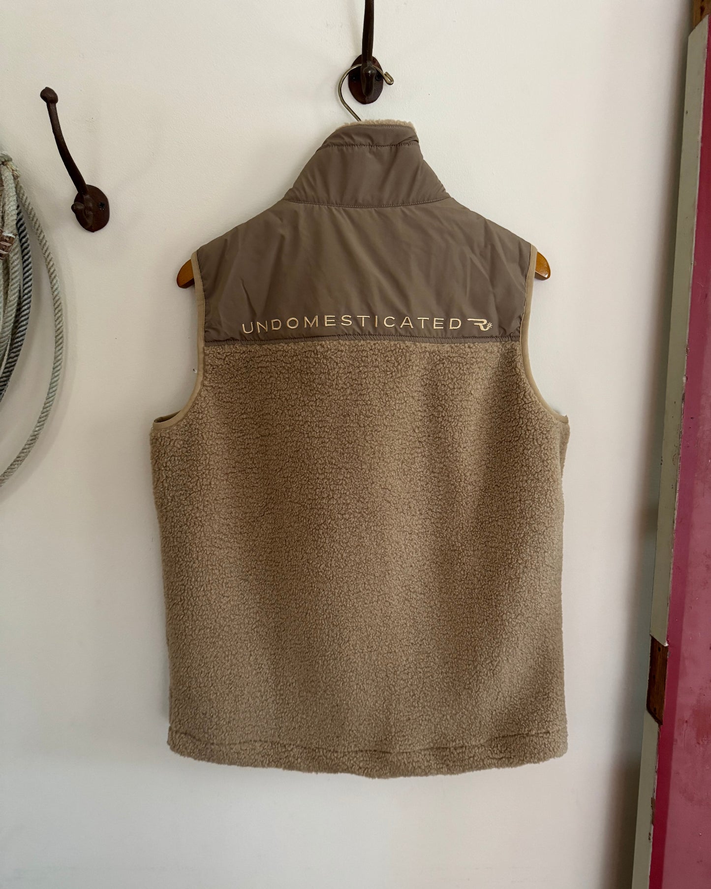 Brown vest with 'UNDOMESTICATED' branding on a white wall.