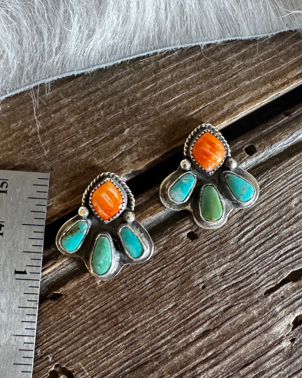 A pair of silver stud earrings with orange cabochon and turquoise inlay, displayed on a rustic wooden surface next to a metal ruler for scale.