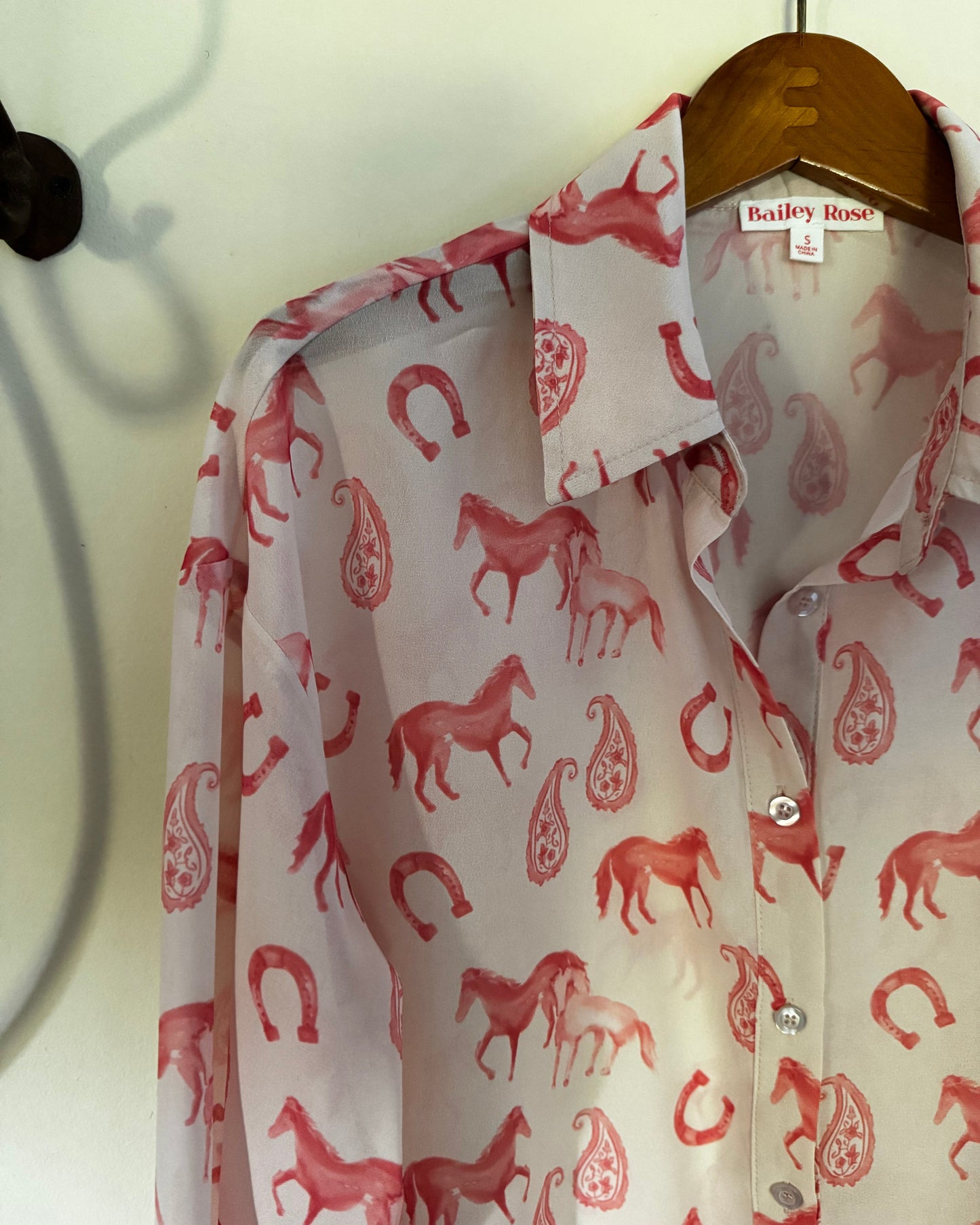 A Bailey Rose button-down shirt with a red and pink horse and paisley pattern, hanging on a wooden hanger against a light wall.